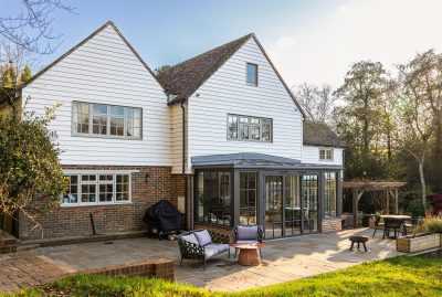 James Wells Architects — A photo of a detached house, which has white wood cladding on the upper storey. There is a black wood extension with Crittal style frams, and a paves